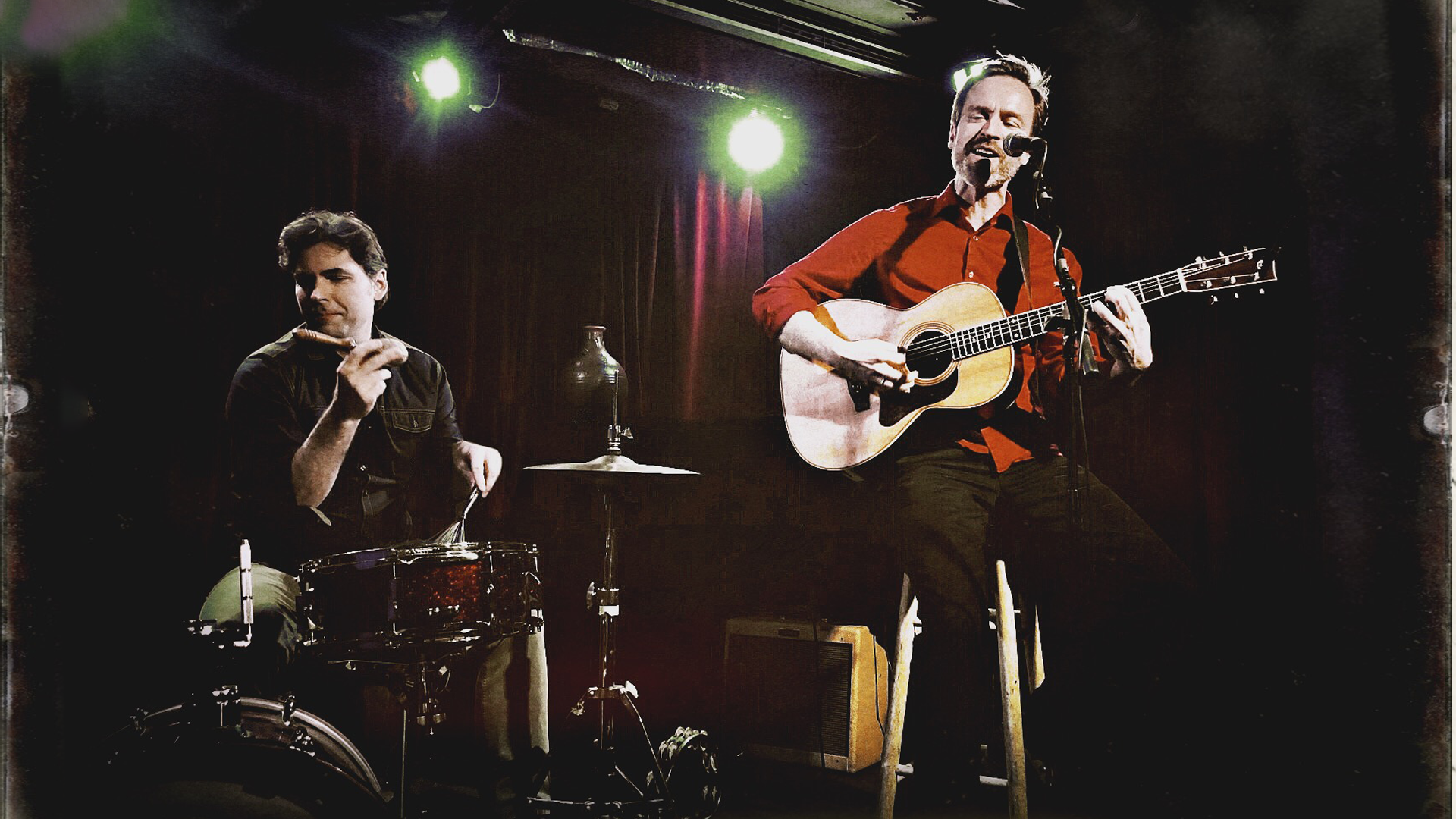 Man with a red shirt playing guitar in front of a man playing drums